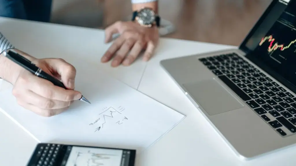 Hand schreibt auf Papier neben Laptop mit Aktienchart bei der Planung des ersten Aktienkaufs