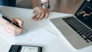 Hand schreibt auf Papier neben Laptop mit Aktienchart bei der Planung des ersten Aktienkaufs